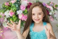 Portrait of a smiling pretty little girl in a turquoise dress holds out a hands near a flowers