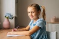 Portrait of smiling pretty primary girl child writing doing homework sitting at home table by window looking at camera. Royalty Free Stock Photo