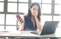 Portrait of smiling pretty young Asian business woman working on laptop at office while holding mobile phone and looking at camera Royalty Free Stock Photo