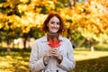 Portrait of smiling pretty young european red-haired lady in raincoat hold red leaf in city park in autumn Royalty Free Stock Photo