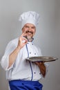 Portrait of a smiling professional chef cook man with a metallic round tray dressed a chef uniform gesturing ok, grey background
