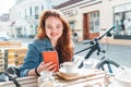 Portrait of smiling red curled long hair caucasian teen girl sitting on a cozy cafe outdoor terrace on the street using the modern Royalty Free Stock Photo