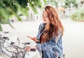 Portrait of smiling red curled long hair caucasian teen girl unlocking bike at Bicycle sharing point using the modern smartphone.