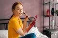 Portrait of smiling redhaired female powdering face with brush looking at camera sitting on bed in bedroom. Side view of Royalty Free Stock Photo