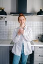 Portrait of smiling redhead woman holding cup of hot beverage in hands on morning at kitchen. Royalty Free Stock Photo