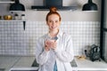 Portrait of smiling redhead woman holding cup of hot beverage in hands on morning at kitchen. Royalty Free Stock Photo