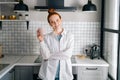 Portrait of smiling redhead woman holding cup of hot beverage in hands on morning at kitchen. Royalty Free Stock Photo