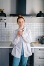 Portrait of smiling redhead woman holding cup of hot beverage in hands on morning at kitchen. Royalty Free Stock Photo