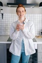 Portrait of smiling redhead woman holding cup of hot beverage in hands on morning at kitchen. Royalty Free Stock Photo