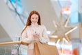 Portrait of smiling redhead young woman using smartphone holding shopping paper bags with purchase standing in hall of Royalty Free Stock Photo