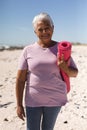 Portrait of smiling retired senior biracial woman holding yoga mat with short white hair at beach Royalty Free Stock Photo