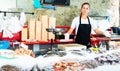 Portrait of smiling saleswoman of fish store standing behind counter with large assortment of fresh seafood