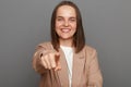 Portrait of smiling satisfied delighted woman with brown hair wearing beige jacket, pointing at camera, choosing you, needs you, Royalty Free Stock Photo