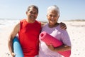 Portrait of smiling senior biracial man with arm around woman holding yoga mats at beach