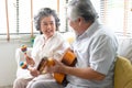 Portrait of smiling senior couple playing guitar singing and her wife holding maracas dancing and sitting on sofa at home.