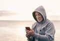 Portrait of a smiling senior man enjoying a cold winter day at sea wearing hood on his head. Looking at mobile phone as the sun Royalty Free Stock Photo