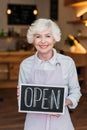 portrait of smiling senior worker in apron holding open chalkboard in hands