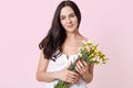 Portrait of smiling sincere young model standing isolated over light pink background, holding colourful spring flowers in hands, Royalty Free Stock Photo