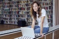 Portrait of smiling smart woman model with opened laptop in a library, bookshelf behind, long hair. Hipster college student lady