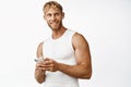 Portrait of smiling strong sportsman with mascules, holding smartphone, using sport app, standing over white background