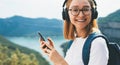 Portrait smiling traveler blonde girl  with hipser glasses and backpack using  mobile phone device and listens music for headphone Royalty Free Stock Photo