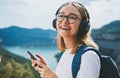 Portrait smiling traveler woman with hipser glasses and backpack holds smartphone device in hands and listens music for headphones Royalty Free Stock Photo