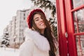 Portrait smiling winter girl with long brunette hair in red knitted hat chilling on street near red telephone box. Snow Royalty Free Stock Photo