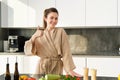 Portrait of smiling woman in bathrobe, shows thumbs up, stands in kitchen, cooking salad, preparing vegetarian dinner