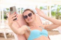 Portrait of smiling woman lying on sun lounger on beach and waving her hand into smartphone Royalty Free Stock Photo