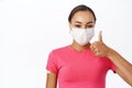 Portrait of smiling woman in medical mask shows right way to wear it, thumbs up, stands in tshirt, has patch on shoulder Royalty Free Stock Photo