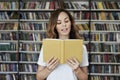 Portrait of smiling woman model reading text with opened book in a library, bookshelf behind, long hair. Hipster college student Royalty Free Stock Photo