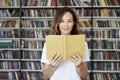 Portrait of smiling woman model reading text with opened book in a library, bookshelf behind, long hair. Hipster college student Royalty Free Stock Photo