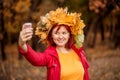 Smiling woman with red hair in crown made of golden leaves snapping picture of herself selfie with smartphone in autumn park Royalty Free Stock Photo