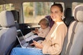 Portrait of smiling woman sitting with her baby daughter in safety chair on backseat of the car, working on laptop, going to Royalty Free Stock Photo