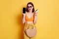 Portrait of smiling woman wearing summer outfit with straw bag doing shopping online, holding smart phone and credit card isolated Royalty Free Stock Photo