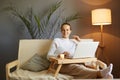 Portrait of smiling woman wearing white sweater with bun hairstyle sitting on cough at home and holding laptop, watching movie on Royalty Free Stock Photo