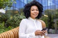 Portrait of a smiling young African American businesswoman sitting on a bench outside an office building, holding a Royalty Free Stock Photo