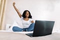 Portrait of smiling young African woman using laptop in bed at home, having video call with her friend, realxing with