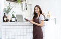 Portrait of a smiling young Asian barista in apron smiling using tablet and looking away on beside coffee machine in counter.