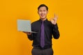 Portrait of smiling young Asian businessman showing okay sign and holding laptop over yellow background Royalty Free Stock Photo