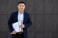 Portrait of a smiling young Asian man in a business suit standing against the wall of an office building, holding Royalty Free Stock Photo