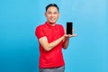 Portrait of smiling young asian man in red shirt showing blank screen mobile phone and looking at camera isolated on blue Royalty Free Stock Photo