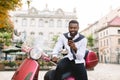 Portrait of smiling young bearded african american guy in smart casual wear using his smartphone, while sitting on the Royalty Free Stock Photo