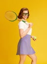 Portrait of smiling, young, beautiful girl in sportswear and glasses, posing with tennis racket and ball against yellow Royalty Free Stock Photo