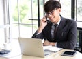 Smiling young businessman talking on his smartphone while he working on his computer, sitting in his office at desk wearing a bla Royalty Free Stock Photo