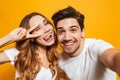 Portrait of smiling young couple man and woman taking selfie photo and showing peace sign, isolated over yellow background Royalty Free Stock Photo