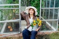 Portrait of smiling young female farmer woman holding watering can with fresh sunflowers bouquet, sits near greenhouse Royalty Free Stock Photo