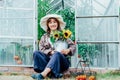 Portrait of smiling young female farmer woman holding watering can with fresh sunflowers bouquet, sitting near greenhouse. Urban Royalty Free Stock Photo