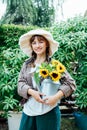 Portrait of smiling young female farmer woman holding watering can with fresh sunflowes bouquet on the green garden Royalty Free Stock Photo