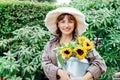 Portrait of smiling young female farmer woman holding watering can with fresh sunflowes bouquet on the green trees Royalty Free Stock Photo